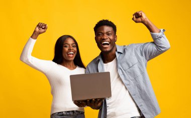Genç bir çift sevinçlerini ve heyecanlarını ifade ediyor. Kutlama için kollarını kaldırarak dizüstü bilgisayar tutuyorlar. Canlı sarı bir arka plana karşı duruyorlar, mutluluklarını ve başarılarını sergiliyorlar..