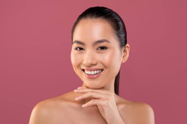 Güzel genç bir Çinli kadın mor bir arka fona üstsüz poz veriyor, kameraya gülümsüyor. Çenesine dokunur, pürüzsüz tenini ve ışıltısını gösterir..