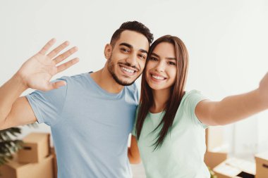 Genç bir çift yeni evlerine taşınmalarını mutlulukla kutluyor. Gülümsüyorlar ve hareket eden kutular ve doğal ışıklarla çevrili selfie için poz veriyorlar..
