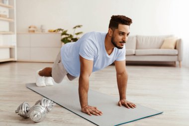 Genç bir adam geniş bir oturma odasında spor minderinin üzerinde şınav çekiyor. Dambıllar yakında ve doğal ışık alanı doldurarak egzersiz için motive edici bir ortam yaratıyor..