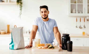 Genç bir adam taze meyve, sebze ve protein takviyeleriyle dolu parlak bir mutfakta duruyor. Besleyici malzemeler kullanarak sağlıklı bir yemek hazırlarken odaklanmış görünüyor..