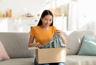 Mutlu kadın rahat oturma odasında kanepesinde oturuyor, neşeyle yeni giysilerini açıyor. Güneş ışığı, internetten alışveriş yapmanın heyecanını yaşayarak son alışverişlerini yaptığı yeri dolduruyor..