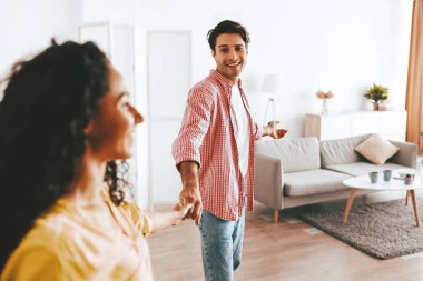 Neşeli bir adam, keyifli bir şekilde gülümseyen bir kadının elini tutar ve sıcak oturma odalarında keyifli bir anı paylaşır. Güneş ışığı pencereden akar ve sıcak bir atmosfer yaratır..