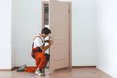 Kırmızı tulumlu yetenekli bir işçi boş bir odada kapı kolunu tamir ederken diz çöküyor. Yeni boyanmış duvarlar ve ahşap döşemeler tadilatta olduğunu gösteriyor..