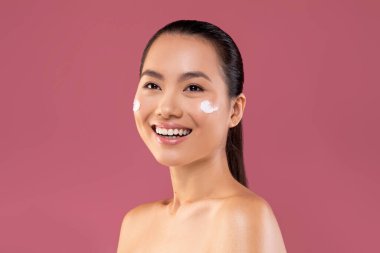 A beautiful young woman smiles while applying moisturizer cream on her cheeks. She is in a purple studio background and enjoying the experience of trying out a new beauty product.