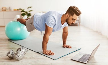 Bir adam, bir dizüstü bilgisayarında antrenman yaparken egzersiz topu kullanarak şınav çeker. Yakınlarda ağırlıklarla aydınlık bir odada, spor salonunu gösteriyor..