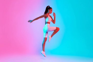 A woman in workout attire performs a dynamic exercise move in a vibrant studio space. She demonstrates strength and agility against a backdrop of pink and blue lighting, embodying energy and focus.