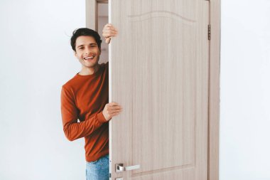 Kırmızı uzun kollu gömlekli neşeli bir adam kısmen açık ahşap bir kapının önünde duruyor. Sıcak bir gülüşü var ve birini karşılamaya hazır görünüyor, aydınlık, basit bir odada.