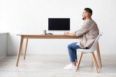 Arap adam parlak bir ev ofisinin ahşap masasında oturuyor, boş ekranlı bir bilgisayar kullanıyor. İşine odaklanırken mutlu görünüyor. Sabah ışığı odayı dolduruyor..