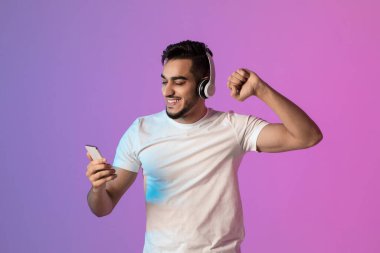 Mutlu genç Arap adam kulaklıkla müzik dinlerken neşeyle dans eder piyangoyu kazandıktan sonra akıllı telefonunu kontrol eder. Heyecanını parlak, renkli bir ortamda ifade ediyor..