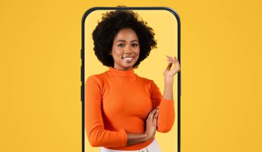 Afro saç stili olan neşeli, genç bir kadın, akıllı bir telefon çerçevesinde, kendinden emin bir şekilde duruyor. Turuncu bir süveter giyiyor ve gülümsüyor, canlı ve çekici bir sahne yaratıyor..