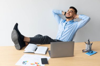 Genç bir Arap masasında sırtüstü oturuyor, gülümsüyor ve dinleniyor. Neşeli bir çalışma ortamında ellerini başının arkasında gevşetirken bacaklarını masanın üzerinde dinlendiriyor..