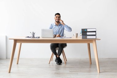 Neşeli bir Arap erkek, şık bir ev ofisinde masasında oturmuş dizüstü bilgisayarla çalışırken cep telefonuyla konuşuyor. Odaklanmış görünüyor ve işini seviyor. Ayar parlak ve minimalistik..