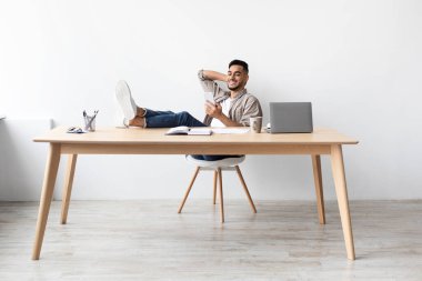 Neşeli bir adam modern bir ofis masasında rahat bir şekilde oturuyor, cep telefonuyla yazı yazıyor. Rahatlamış çalışma alanının tadını çıkarırken, ayaklarını masaya dayayıp gülümsüyor..