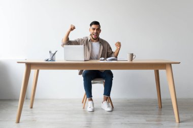 Neşeli bir Arap adam, ofis masasında dizüstü bilgisayar kullanarak oturuyor. Kutlamak için kollarını kaldırır ve yumruklarını sıkar. Son başarısında heyecanını ve mutluluğunu ifade eder..