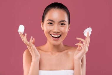 Havluya sarılmış Koreli genç bir kadın yüz bakımının keyfini çıkarıyor. Elinde pamuk pedler ve mor bir stüdyo arka planında gülücükler tutuyor, cilt aktivitesini vurguluyor..