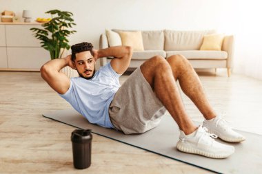 Genç bir adam, iyi aydınlatılmış bir oturma odasında yoga minderinde mekik çekerek egzersiz yapıyor. Günlük spor kıyafetleri giyiyor ve formunu korumaya odaklanıyor..