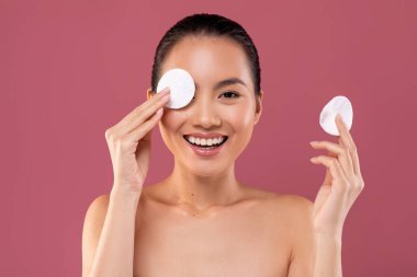 Genç Japon kadın gülümser ve makyaj malzemelerini çıkarırken gözünü pamuk pedlerle kaplayarak poz verir. Mor renkli arka plan neşesini arttırıyor..