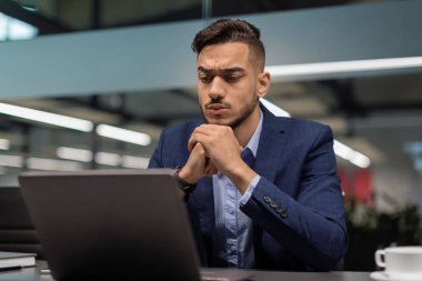 Gösterişli bir ofiste, düşünceli, genç, orta doğulu bir iş adamının yakın çekimi. Bir dizüstü bilgisayar ekranına odaklanmış, pazarlama araştırmaları yürütüyor ve yeni iş fırsatları keşfediyor..