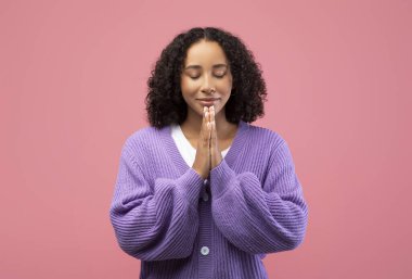 In a bright pink studio, a young African American woman with curly hair gently clasps her hands together and closes her eyes in a moment of prayer, seeking peace and blessings.
