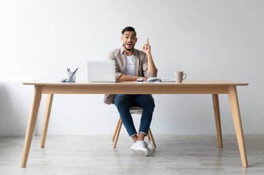 Genç bir adam bir dizüstü bilgisayarla ahşap bir masada oturuyor, şaşkın şaşkın bakıyor ve bir çözüm bulmuş gibi parmağını kaldırıyor. Ofisin temiz, minimalist bir tasarımı var..