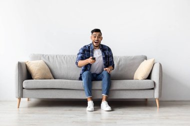 Neşeli genç adam sıcak bir oturma odasında kanepeye oturur, elinde uzaktan kumandayla büyüleyici bir şovu hevesle seyreder. İfadesi boş zamanlarında saf heyecan ve mutluluk gösteriyor..