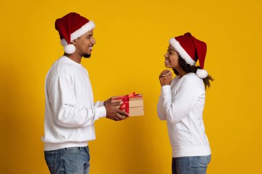 Noel Baba şapkası takan iki insan birbirlerine bir hediye verirken gülümserler. Arka plan parlak sarı, tatil sezonu için şenlikli bir ortam yaratıyor..