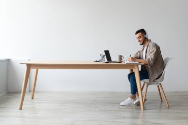Neşeli genç bir Arap, bir masada kulaklık takarak oturuyor. İnternette çalışırken bir dizüstü bilgisayar kullanıyor ve bir deftere yazıyor. Doğal ışık sessiz boşluğu doldurur..