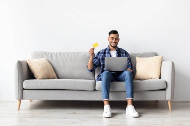 Genç adam kanepede oturuyor, dizüstü bilgisayar kullanırken gülümsüyor ve kredi kartı tutuyor. Oturma odasında online alışveriş yapıyor..