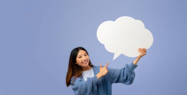 A young woman smiles while pointing at a large, blank speech bubble shape. She stands in front of a soft blue background, expressing excitement or enthusiasm. Her casual outfit adds a friendly tone.