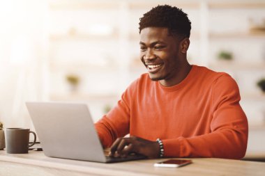 Genç bir adam parlak ve rahat bir çalışma alanında ahşap bir masada oturuyor. Dizüstü bilgisayarında yazarken gülümsüyor, rahat ve neşeli bir atmosfer yaratıyor..