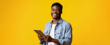 Neşeli bir adam elinde tabletle ayakta duruyor, açık tişört giymiş, parlak sarı bir arka plana karşı. Şu anda meşgul ve mutlu görünüyor..