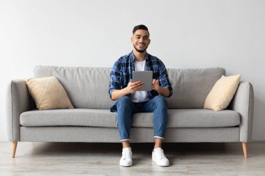 Genç bir Arap, oturma odasındaki rahat bir koltukta oturur ve dijital tablet kullanırken kameraya gülümser. Rahat ve meşgul görünüyor, modern bir ev ortamında internette geziniyor..