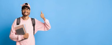 Mutlu bir genç adam parlak mavi bir arka planın önünde duruyor. Elinde kitap ve sırt çantası tutarken pembe bir sweatshirt ve gözlük takıyor ve okulda başarıyı simgelemek için neşe içinde başparmağını kaldırıyor..