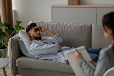 A young arab male patient is lying on a couch in a psychologists office, appearing distressed and seeking help for his depression. The counselor is listening attentively nearby.