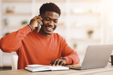 İyi ışıklandırılmış bir odada telefonla konuşurken kırmızı kazak giyen genç bir adam gülümsüyor. Dizüstü bilgisayarının yanına not alıyor, işine odaklandığını ve meşgul olduğunu gösteriyor..