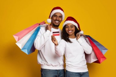 Neşeli bir çift, parlak sarı bir arka plana karşı duruyor, Noel Baba şapkaları takıyor ve alışveriş torbaları tutuyor. Bir kişi kredi kartı gösterirken gülümserler. Tatil alışverişine hazırdırlar..