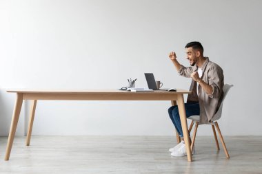 Bir Arap, bir ofis masasında oturmuş başarıyı kutluyor. Heyecanla yumruklarını sallıyor ve dizüstü bilgisayarına bakarken evet diye bağırıyor..