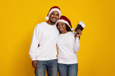 Neşeli bir çift, her ikisi de Noel Baba şapkası takan parlak sarı bir arka plana karşı birlikte duruyor. Gülümsüyorlar ve tatil maceraları için heyecanlı seyahat belgeleri tutuyorlar..