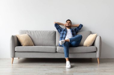 Neşeli Orta Doğulu adam oturma odasındaki rahat bir kanepede dinleniyor. Gülümseyerek arkasına yaslanıyor, boş zamanlarının tadını çıkarıyor ve işten uzaklaşıyor, rahat ve mutlu hissediyor..