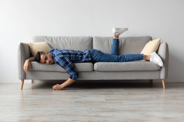 Genç bir Orta Doğulu erkek, gri bir kanepeye yüzüstü uzanmış uzun bir çalışma gününden sonra yorgun ve bitkin bir halde. Evde, biraz dinlenmek ve dinlenmek istiyor..