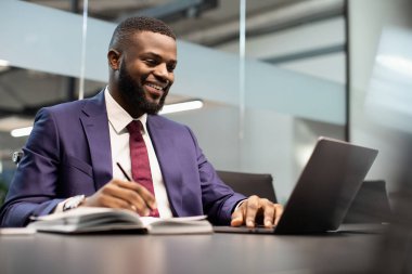 Ofiste çalışan gülümseyen Afrikalı Amerikalı iş adamı, masada oturuyor, modern bilgisayar kullanıyor ve notlar alıyor, İnternet 'teki iş konferansına katılıyor,