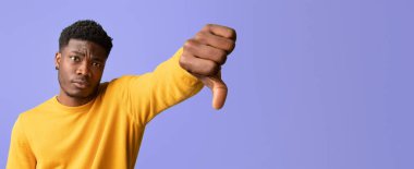 Kısa siyah saçlı genç bir adam baş parmak aşağı hareket ederken sarı bir kazak giyer. Arka plan pürüzsüz bir mor renkte. Onaylamama ifadesini vurguluyor..