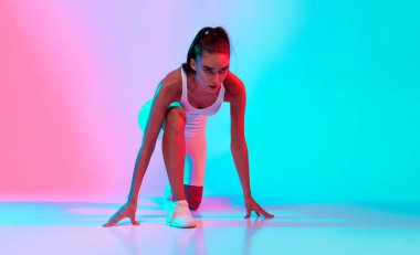 Dişi bir atlet başlangıç pozisyonunda çömelmiş, koşmaya hazır. Arka plan mavi ve pembe renklerle aydınlatılmış, enerjik ve motive edici bir atmosfer yaratıyor..