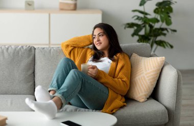 Genç bir Arap kadın bir fincan kahve tutarak rahat bir kanepede dinleniyor. Düşünceli bir şekilde yan tarafa bakıyor, evde huzurlu bir hafta sonu sabahının sükunetini kucaklıyor..