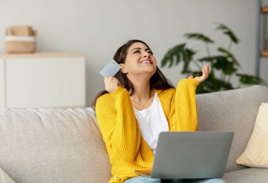 Mutlu bir Arap kadın kanepesinde dizüstü bilgisayar kullanarak ve kredi kartı tutarak oturuyor. Büyük bir satıştan faydalanırken online alışveriş için heyecanlanıyor. Doğal ışık odayı dolduruyor..