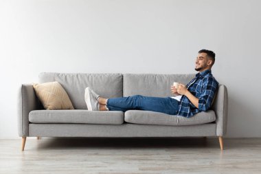 Neşeli bir Arap, oturma odasındaki rahat bir koltuğa rahatça oturur. Evde dinlenme molası sırasında kahvesini içiyor, huzurlu bir hafta sonu anını kucaklıyor..