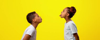 İki çocuk birbirine yakın duruyor, birbirlerine aptalca suratlar yapıyorlar. Bir çocuk dilini uzatırken diğeri umutla bakıyor. Parlak sarı arka plan canlı bir his veriyor..