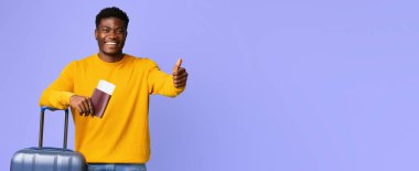 Genç bir adam bir bavulun yanında dururken gülümsüyor ve başparmağını kaldırıyor. Bir elinde pasaportu var, yaklaşan yolculuğunun beklentilerini ifade ediyor..