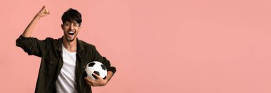 Gözlüklü genç bir adam bir futbol topu tutarken sevinçle kolunu kaldırıyor. Gülümsüyor ve günlük kıyafetler giyiyor. Arkaplan koyu pembe renktedir..